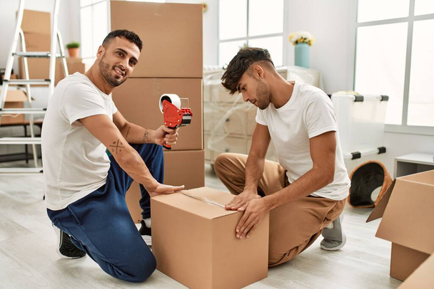 Dos hombres hispanos pareja sonriendo caja de cartón de embalaje seguro en nuevo hogar - Foto, imagen