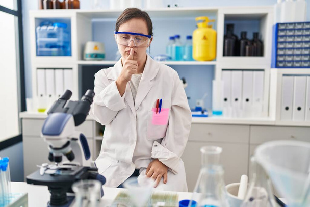 Hispanic girl with down syndrome working at scientist laboratory asking to be quiet with finger on lips. silence and secret concept.  - Photo, Image