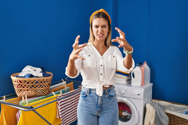 Young blonde woman at laundry room shouting frustrated with rage, hands trying to strangle, yelling mad  - Photo, image