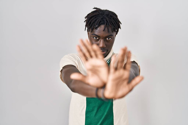 Αφρικανός άνδρας με dreadlocks στέκεται πάνω από απομονωμένη έκφραση απόρριψης υποβάθρου διασχίζοντας τα χέρια και τις παλάμες κάνει αρνητικό σημάδι, θυμωμένος πρόσωπο  - Φωτογραφία, εικόνα