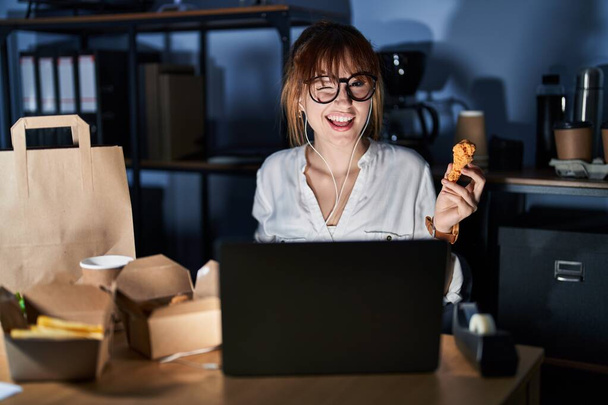Fiatal gyönyörű nő dolgozik számítógép laptop és étkezési szállítás élelmiszer kacsintás néz a kamera szexi kifejezés, vidám és boldog arc.  - Fotó, kép
