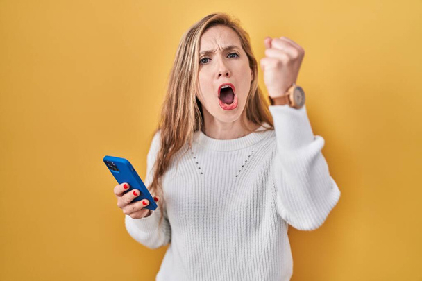 Jonge blonde vrouw met behulp van smartphone typen bericht boos en gek verhogen vuist gefrustreerd en woedend terwijl schreeuwen van woede. woede en agressief concept.  - Foto, afbeelding