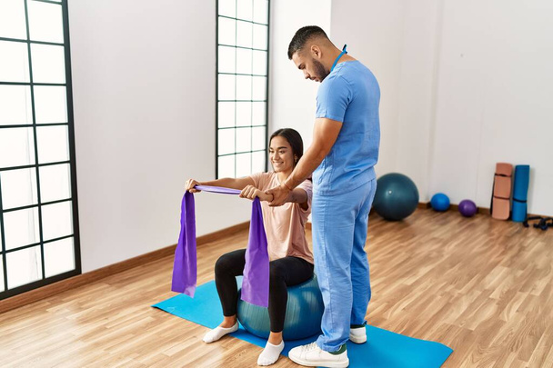 Homem e mulher latinos vestindo uniforme fisioterapeuta tendo sessão de reabilitação usando bola de ajuste e banda elástica no centro de reabilitação - Foto, Imagem