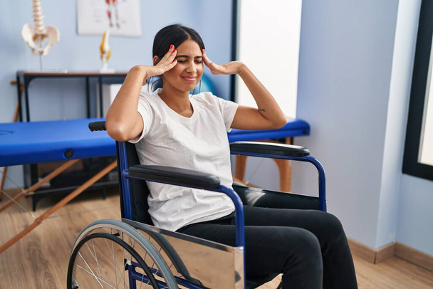 Young hispanic woman sitting on wheelchair at physiotherapy clinic with hand on head, headache because stress. suffering migraine.  - Photo, Image
