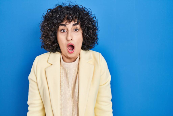 Young brunette woman with curly hair standing over blue background afraid and shocked with surprise and amazed expression, fear and excited face.  - Photo, Image