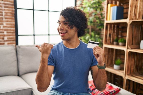 Homme hispanique avec les cheveux bouclés tenant carte de crédit pointant pouce vers le côté souriant heureux avec la bouche ouverte  - Photo, image