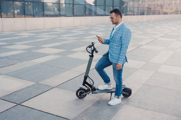 Портрет бизнесмена в синем костюме со смартфоном и скутером на открытом воздухе в городе. - Фото, изображение