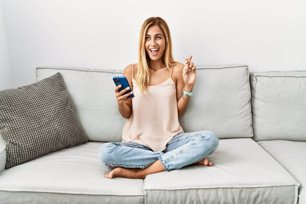 Blond mooie jonge vrouw zitten op de bank thuis met behulp van smartphone gebaren vinger gekruist glimlachen met hoop en ogen dicht. geluk en bijgelovig concept.  - Foto, afbeelding