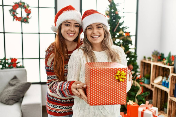 Woman couple hugging each other holding gift standing by christmas tree at home - Foto, Imagem