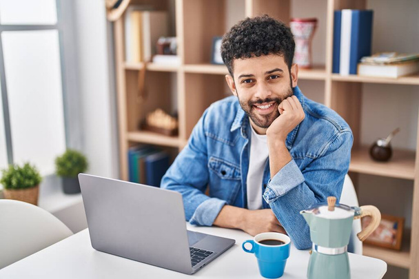 Genç Arap adam dizüstü bilgisayar kullanıyor. Evdeki masada kahve içiyor. - Fotoğraf, Görsel