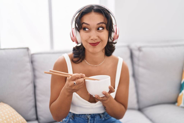 Joven mujer hispana comiendo comida asiática usando palillos sonriendo mirando hacia un lado y mirando hacia otro pensando.  - Foto, imagen