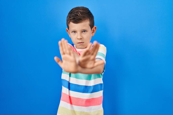 Молода біла дитина, що стоїть на синьому фоні, відмовляється перетинати руки і долоні, роблячи негативний знак, сердите обличчя
  - Фото, зображення
