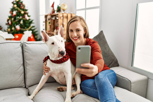 Junge Kaukasierin macht Selfie mit dem Smartphone, sitzt mit Hund am Weihnachtsbaum zu Hause - Foto, Bild