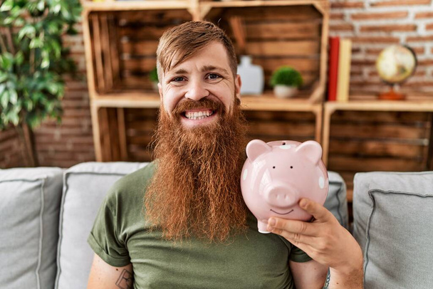 Redhead man holding piggy bank looking positive and happy standing and smiling with a confident smile showing teeth  - Photo, Image