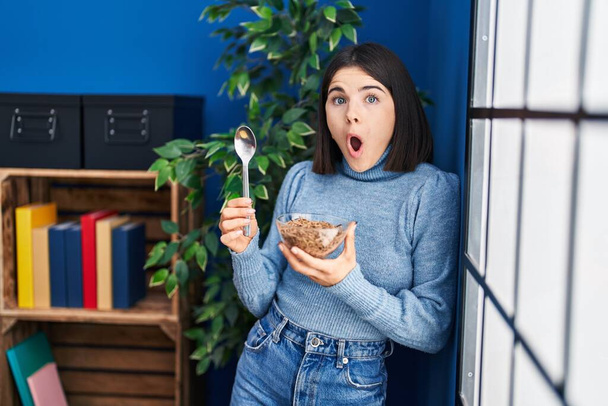 Mujer hispana joven comiendo cereales integrales saludables con cuchara asustada y sorprendida con expresión sorpresa y asombro, miedo y cara excitada.  - Foto, imagen
