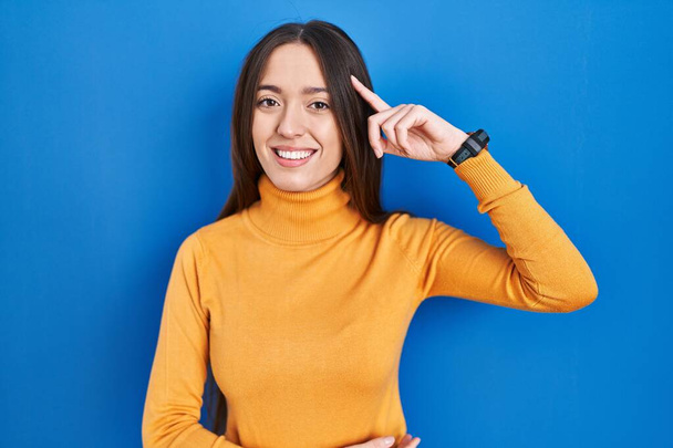 Jeune femme brune debout sur fond bleu souriant pointant vers la tête avec un doigt, bonne idée ou pensée, bonne mémoire  - Photo, image
