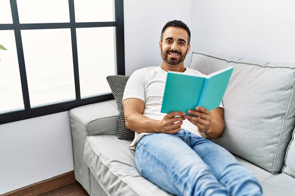 Молодой латиноамериканец улыбается уверенно читая книгу дома - Фото, изображение