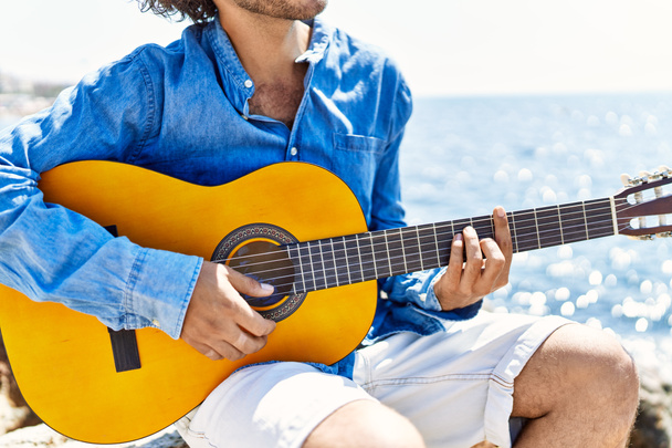 Klasik gitar çalan bir adam plajda kayanın üzerinde oturuyor.. - Fotoğraf, Görsel