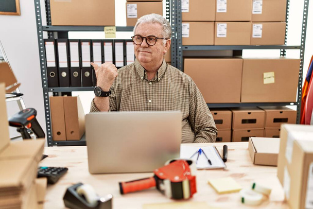 Senior caucasian man working at small business ecommerce with laptop smiling with happy face looking and pointing to the side with thumb up.  - Photo, image