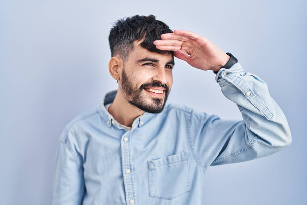 Joven hombre hispano con barba de pie sobre fondo azul muy feliz y sonriente mirando lejos con la mano sobre la cabeza. concepto de búsqueda.  - Foto, imagen