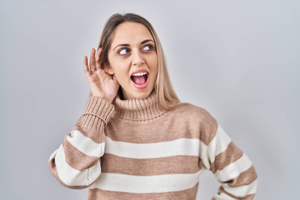Jonge blonde vrouw draagt coltrui over geïsoleerde achtergrond glimlachend met hand op oor luisterend naar geruchten of roddels. doofheid.  - Foto, afbeelding