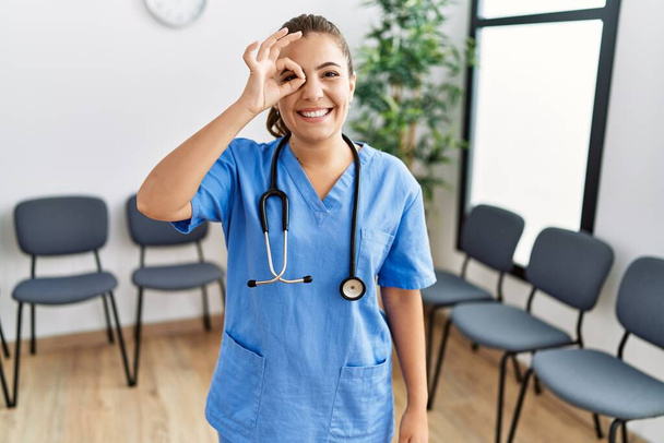 Young brunette doctor woman at waiting room smiling happy doing ok sign with hand on eye looking through fingers  - Photo, Image
