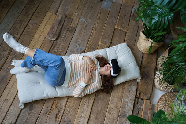 Ηρεμία γυναίκα σε VR ακουστικά βρίσκεται στο ξύλινο πάτωμα στο σπίτι σε surround πράσινα εξωτικά φυτά, εξερεύνηση επαυξημένης πραγματικότητας, παίζοντας διαδραστικά παιχνίδια βίντεο, παρακολουθώντας σειρά. Εικονική έννοια ψυχαγωγίας. - Φωτογραφία, εικόνα