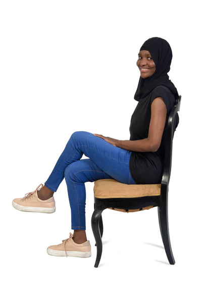 πλαϊνή άποψη μιας γυναίκας με casual ρούχα και μπούρκα κάθεται στην καρέκλα και σταυρωτά πόδια και κοιτάζοντας κάμερα σε λευκό φόντο - Φωτογραφία, εικόνα