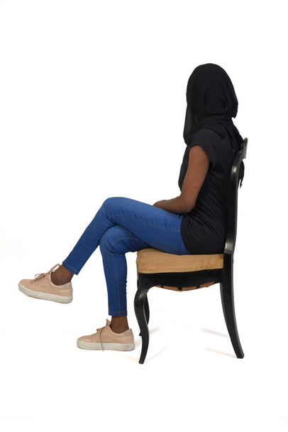 πλαϊνή άποψη μιας γυναίκας με casual ρούχα και μπούρκα κάθεται στην καρέκλα cross-leggedand lookiong φόντο σε λευκό φόντο - Φωτογραφία, εικόνα