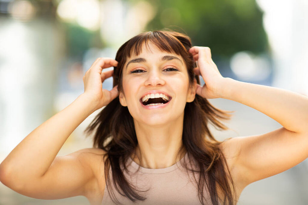 Close up portrait of laughing young woman with hands in hair looking at camera outside - Фото, изображение