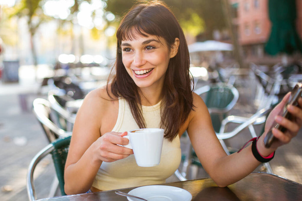 Portrét krásné mladé ženy sedící v venkovní kavárně s telefonem pití šálek kávy - Fotografie, Obrázek
