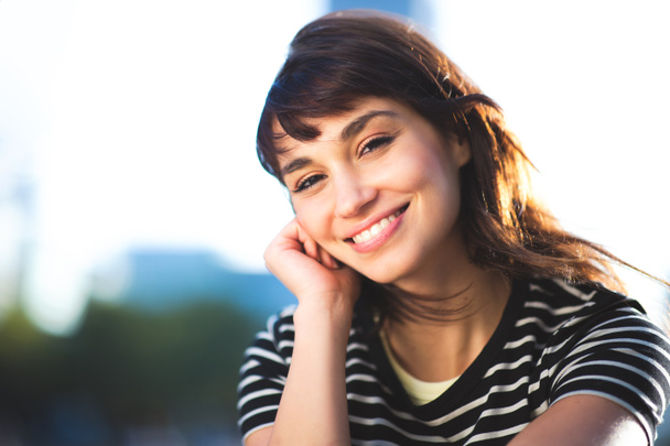 Close up portrait of cheerful young female sitting outside in the city with hand on chin looking at camera and smiling - Photo, Image