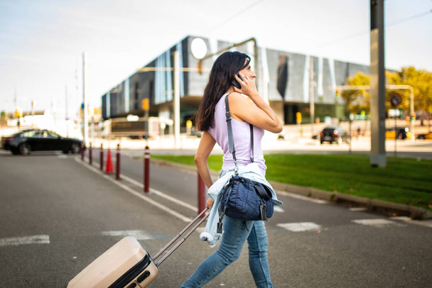 Profiili onnellinen valkoihoinen nainen vetämällä liikkuva matkalaukku ja kuljettaa käsilaukku puhuessaan matkapuhelin ulkona kaupungissa - Valokuva, kuva