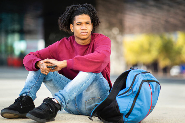 Afričan Američan s mobilním telefonem a batohem sedí venku na ulici - Fotografie, Obrázek