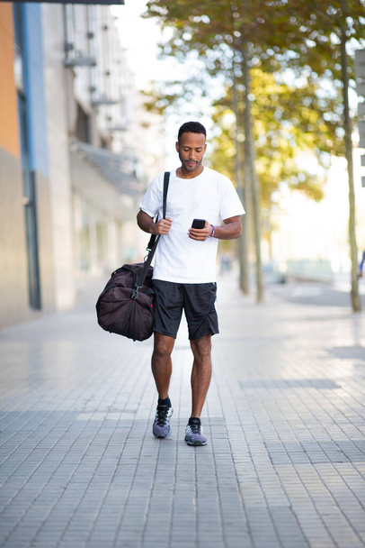 Мужчина носит спортивную сумку на улице, используя мобильный телефон - Фото, изображение