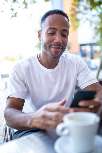 Smiling african american man sitting at a coffee shop and reading text messaging on his mobile phone - Photo, Image