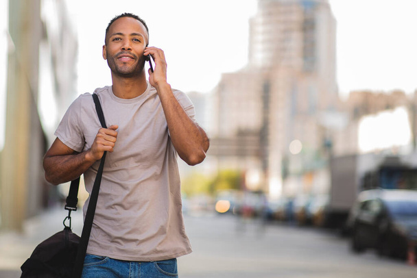 Afroamerikaner telefoniert mit Handy, während er Tasche im Freien in der Stadt trägt - Foto, Bild