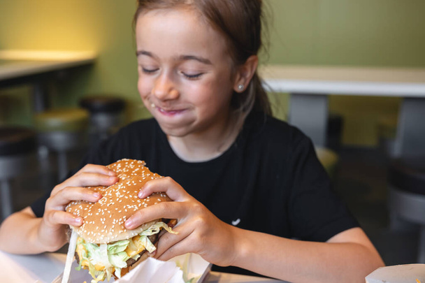 A little girl eats a big appetizing burger in a cafe, close-up. - Foto, Imagen