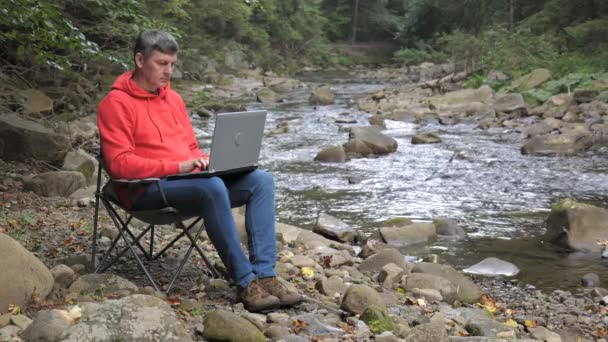 Ένας άντρας δουλεύει σε ένα λάπτοπ κοντά σε ένα ρέμα στη φύση. Έννοια της freelancing, digital nomad ή απομακρυσμένο γραφείο. 4K - Πλάνα, βίντεο