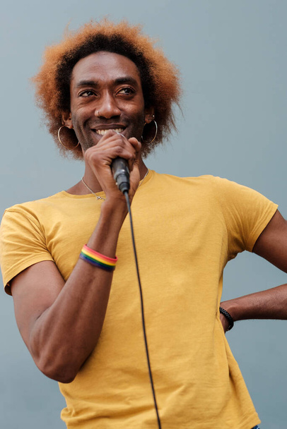 Портрет улыбающегося молодого человека, поющего на улице с микрофоном в руках. Носит браслет lgtbq, вьющиеся волосы. - Фото, изображение