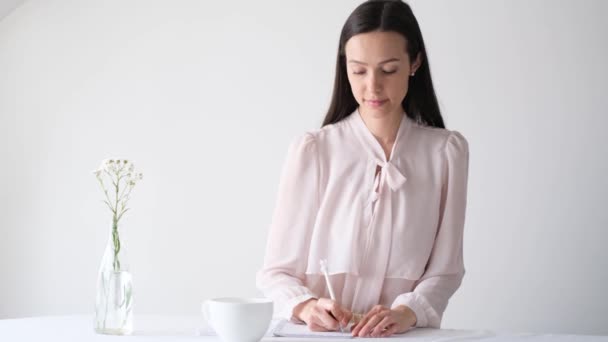  A girl in a shirt sits at a table and writes in a diary in a room. Jotting information, create to-do list, planning concept. - Materiał filmowy, wideo