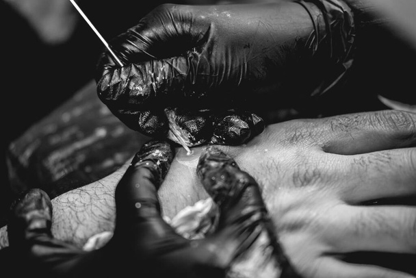 Detalle de las manos del tatuador con guantes negros haciendo un nuevo tatuaje con tinte blanco de yin yang en la mano de un tipo con la técnica tradicional japonesa de "empujar la mano" (en blanco y negro)) - Foto, Imagen
