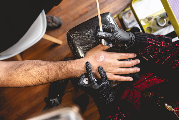 Detail van handen van tatoeëerder met zwarte handschoenen maken van een nieuwe tatoeage met witte tint van yin yang in de hand van een man met traditionele Japanse 'hand porren' techniek - Foto, afbeelding
