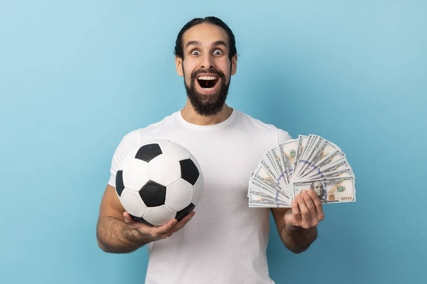 Portrait of man with beard wearing white T-shirt holding soccer ball and lots dollar banknotes, looking camera with amazed face, betting and winning. Indoor studio shot isolated on blue background. - 写真・画像