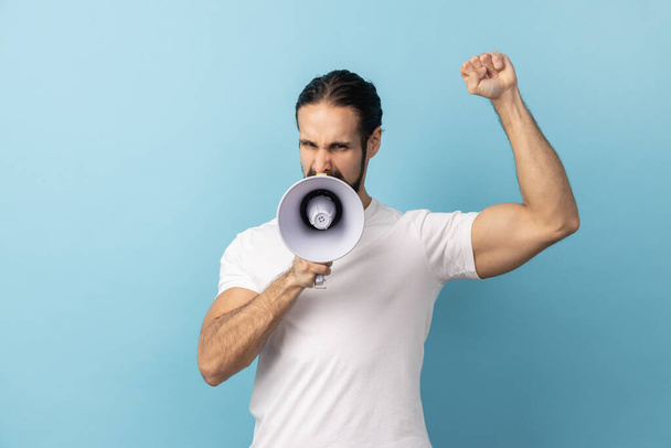 Portrait of angry man with beard wearing white T-shirt loudly screaming at megaphone, making announce, protesting, wants to be heard. Indoor studio shot isolated on blue background. - Photo, image