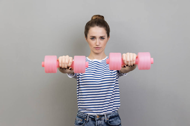 Portrait of serious concentrated woman wearing striped T-shirt holding out pink dumbbells to camera, working out alone. Indoor studio shot isolated on gray background. - Foto, Imagen