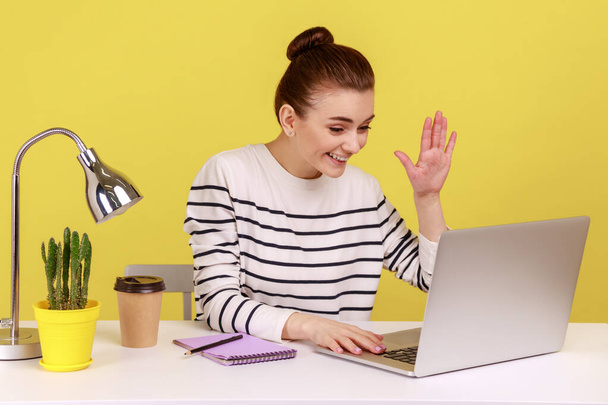 Friendly woman office manager waving hand to laptop screen, talking on video call with friend, online conference at home office. Indoor studio studio shot isolated on yellow background. - Photo, Image