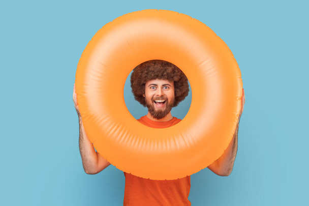 Portrait of man with Afro hairstyle with happy facial expression looking trough bright orange rubber ring, looking at camera, enjoying his vacation. Indoor studio shot isolated on blue background. - Photo, Image