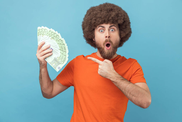 Portrait of surprised amazed man with Afro hairstyle wearing orange T-shirt holding euro banknotes, being shocked to win lottery. Indoor studio shot isolated on blue background. - Foto, afbeelding