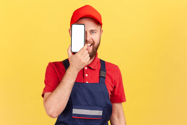 Portrait of happy smiling joyful bearded worker man standing and covering eye with cell phone with blank screen for promotional text. Indoor studio shot isolated on yellow background. - Φωτογραφία, εικόνα